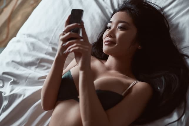 an attractive young asian woman in black underwear is using her phone while looking on her bed and smiling